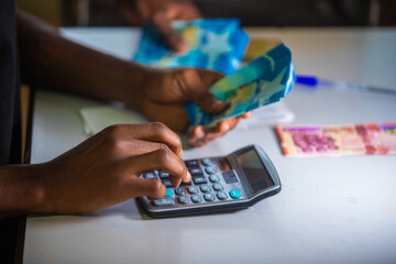 Dark skinned African person working on desk holding Ghanaian cedi notes in hand and pressing punching calculator with some notes on table. Top view accounting concept