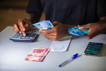 Dark skinned African person working on desk writing in notebook and holding Ghanaian cedi notes in hand with some notes on table. Top view accounting concept