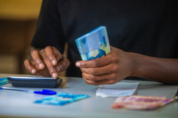 vertical shot of Dark skinned African person working on desk holding counting Ghanaian cedi notes money with some notes on table. accounting concept. 
