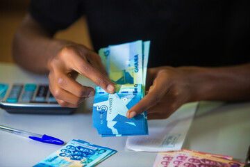 Dark skinned African person working on desk holding and counting Ghanaian cedi notes in hand. Top view accounting concept