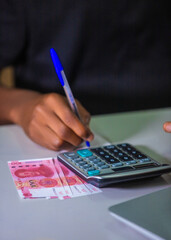 Virtical photo of Dark skinned African person hand fingers holding blue pen writting with calculator and one hundred Chinese renminbi Yuan notes on white table desk
