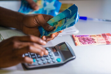 Dark skinned African person working on desk holding Ghanaian cedi notes in hand and pressing punching calculator with some notes on table. Top view accounting concept