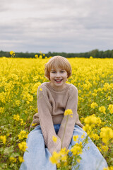Fototapeta premium Front view of 10 year old Caucasian girl with blonde short hair in beige sweater and jeans sitting in yellow rapeseed field, smiling, look at camera. Concept childhood, joy, freedom, copy space.
