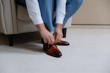 Man tying shoelaces on classic brown shoes, dressed in elegant blue pants and white shirt
