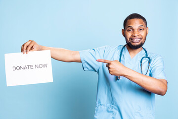 African american nurse advertising the message to donate blood, holding a paper and urging people to help in studio. Medical assistant in workwear standing against blue background.