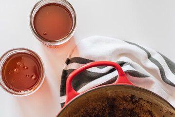 Homemade ghee process in bright natural light, golden clarified butter in glass mason jars beside a red cast iron pot and striped tea towel, high contrast kitchen scene with warm shadows and bright