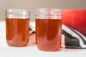 Homemade ghee process in bright natural light, golden clarified butter in glass mason jars beside a red cast iron pot and striped tea towel, high contrast kitchen scene with warm shadows and bright