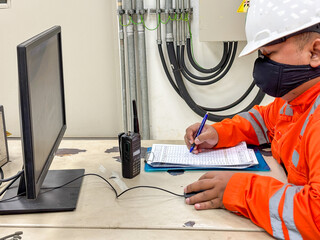 Engineer inspecting energy distribution systems wearing safety gear and mask entering data into report during COVID-19 pandemic while monitoring operations at plant workstation