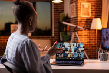 Black woman joining video call with team, working from home on laptop after sunset. Focused female entrepreneur in virtual meeting, handling evening business discussions with remote colleagues.