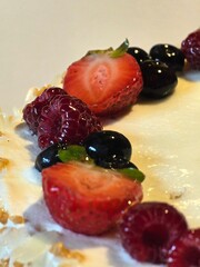 A close-up view of a dessert, cake, decorated with a row of fresh strawberries, raspberries, and blueberries on white frosting.
