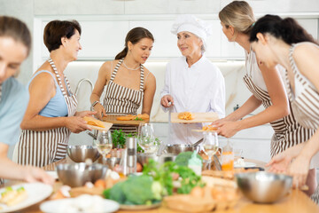 Mature female cook holding cutting board with raw chicken breast in her hands teaching members of cooking course