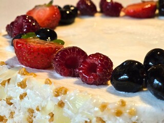 A close-up shot of a delicious-looking cake topped with fresh strawberries, raspberries, and blueberries. The side of the cake is covered in white chocolate flakes.