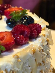 A close-up shot of a delicious-looking cake topped with fresh strawberries, raspberries, and blueberries. The side of the cake is covered in white chocolate flakes.