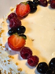 A close-up view of a dessert, cake, decorated with a row of fresh strawberries, raspberries, and blueberries on white frosting.
