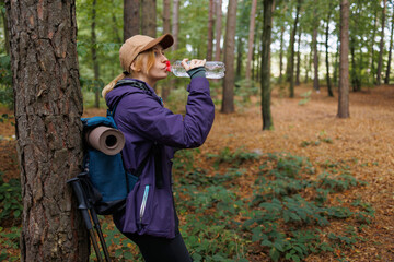 Woman resting in forest, drinking water after hike with backpack and trekking poles