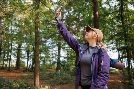 Smiling female hiker raising phone in forest to search for signal during hiking trip