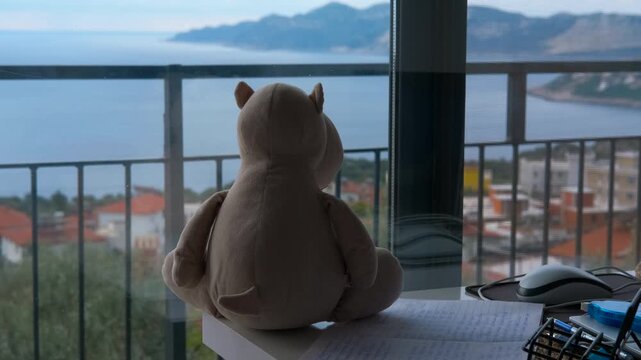 Dreaming about traveling. Plush hippopotamus contemplating inspiring seascape from balcony. Soft plush hippopotamus sitting quietly on wooden desk, gazing longingly through balcony window at seascape