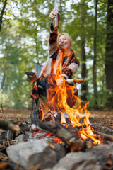 Happy woman raising beer and roasting sausage over campfire flames outdoors