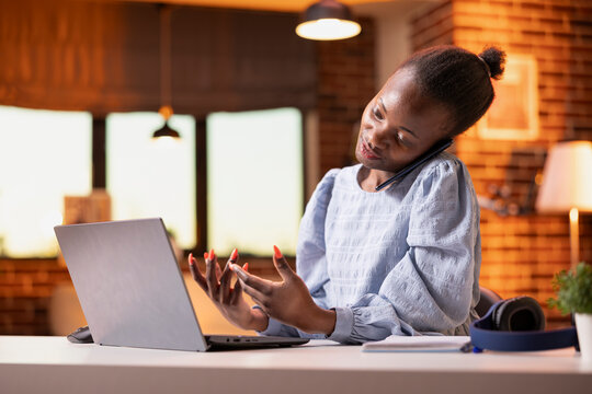 African american female entrepreneur multitasking between laptop and phone call. Black businesswoman holds cellphone to ear while reading information on laptop during client discussion.
