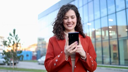 Young professional businesswoman with curly hair texting on her smartphone. Happy female entrepreneur checking social media on the go - Powered by Adobe
