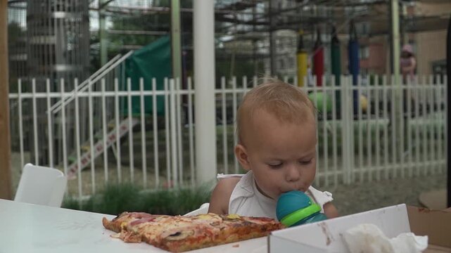 A woman gives a baby girl a blue sippy cup with water from a straw while they dine on pizza in a pizzeria on a hot summer day. A huge slice sits on the table.