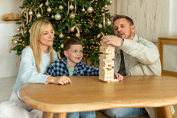 Happy family plays jenga with child on Christmas, enjoying board game and removing a wooden blocks from the unstable tower. Satisfied boy developing fine motor skills. High quality photo