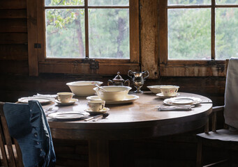 Rustic cabin interior with a round wooden table set with elegant tableware, including bowls, tea set, and plates, by the window