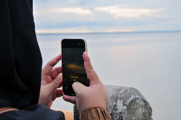 A person holds up a smartphone to photograph the tranquil sunset over a wide river. The camera screen displays the scene, highlighting the modern way of documenting natural beauty.