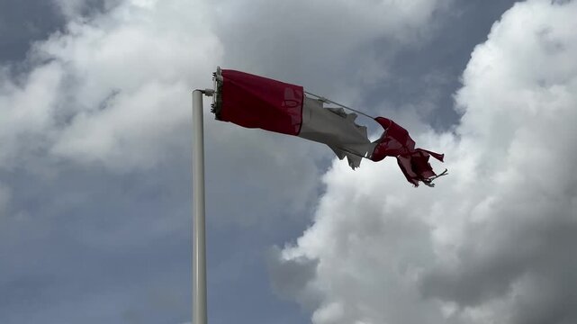 A tattered, torn, and weathered windsock blows in a strong wind against a cloudy sky