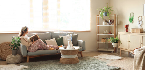 Young witch with spell book sitting on sofa at home