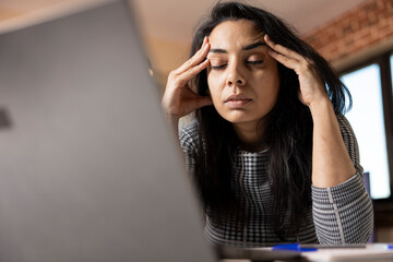 Fatigued indian woman seated at desk with eyes closed, taking break and massaging her temples. Female business consultant battling headache and struggling to stay focused while working from home.