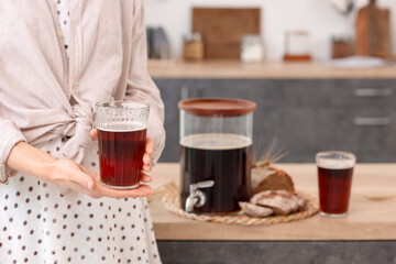 Young woman with glasses of tasty kvass with bread slices and spikelets in kitchen, closeup
