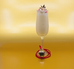 Tall glass of eggnog with whipped cream, star anise and peppermint pieces on top. A bell with a red cord is wrapped around the bottom of the glass. The background is a gold metallic color.