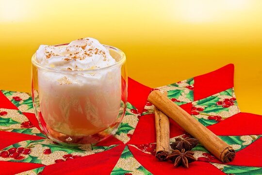 Glass of eggnog with whipped cream and cinnamon on top. The glass sits on a Christmas themed cloth. Cinnamon sticks and star anise are also on the cloth. There is a gold metallic background.