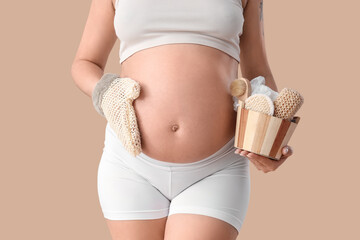 Beautiful young pregnant woman with massage mitten and brushes on beige background, closeup
