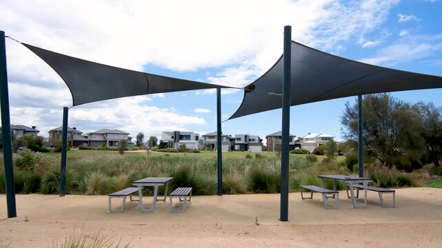 Public picnic area with shade sails, tables, and benches in suburban parkland in Werribee South, Australia, with modern houses in the background. Concept of community facilities, outdoor leisure