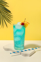 Glass of tasty Blue Lagoon cocktail garnished with cherry and palm leaf on white table against yellow background
