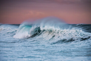 Powerful wave breaking at Bronte Beach, Sydney, Australia, during sunset. Soft evening light colors the sky as ocean spray rises dramatically.