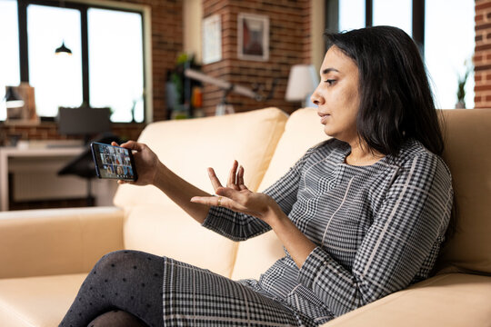 Indian female executive seated on sofa with cellphone, speaking remotely with diverse employees on video call. Professional woman conducting impromptu virtual team meeting on mobile phone from home.