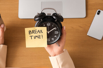 Female hand holding paper with text BREAK TIME and alarm clock in office, top view