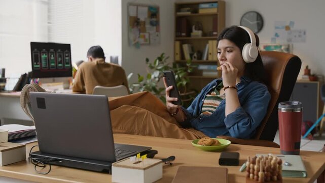 Young female programmer sitting in office chair, putting his feet on table. She taking oatmeal cookie, biting it while scrolling social network feed