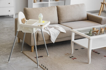Baby highchair with toys and bottle of milk near sofa in living room interior