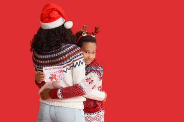 African-American mother and her daughter with Christmas greeting card hugging on red background