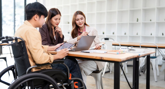 A diverse team of professionals, including a man in a wheelchair, collaborate and discuss project details on a laptop at a modern office.