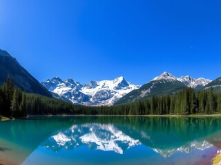 Tranquil alpine lake reflects snow-capped mountains and pine forest under a clear blue sky