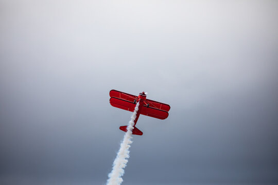 A restored World War II era red biplane airplane performing aerobatic maneuvers in the sky during an air show event, leaving a trail of smoke against a cloudy background in Hillsboro, Oregon, USA