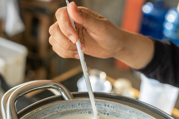 A hand uses a thermometer to check the temperature of fresh cow milk in a metal pot during cooking preparation in a kitchen.