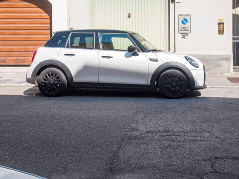 Mini Cooper F55 five door hatchback in pepper white with a black roof parked on a sunlit European street beside garage doors and a passo carrabile sign