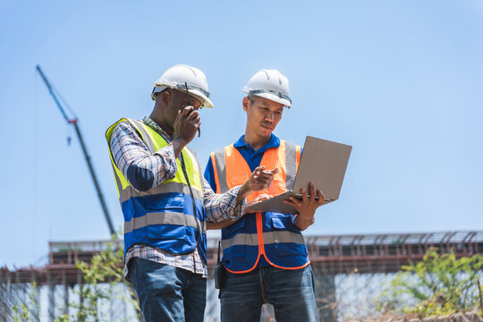 Construction Engineers Using a Laptop and Walkie-Talkie on Site, Multicultural Team of Foreman Worker Planning with Digital Tools, Professional Project Managers at a Construction Site Reviewing Data - Powered by Adobe