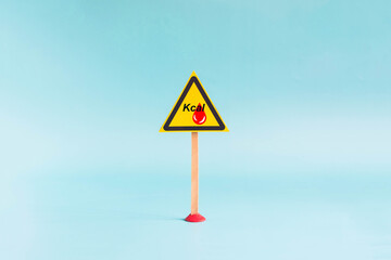 The "calories" warning sign in the form of a yellow triangle on a stand, close-up on a light background. The concept of a warning sign about high-calorie foods